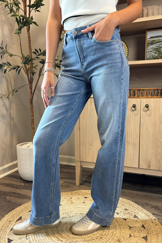 A close-up view of a woman wearing light-wash high-waisted jeans paired with a white sleeveless knit top. She stands indoors on a woven circular rug, wearing white sneakers, with part of a potted plant and wooden shelving visible in the background.