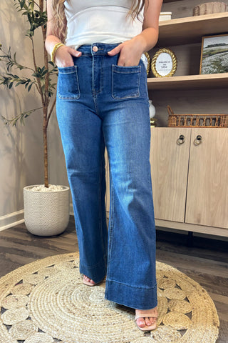 Close-up of a woman wearing high-waisted dark blue wide-leg jeans with large front patch pockets. She pairs them with a white ribbed tank top and nude open-toe block heels, standing on a woven circular rug with part of a potted plant and wooden shelving in the background.
