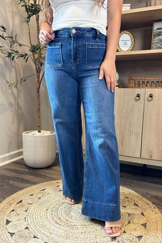 Close-up of a woman wearing high-waisted dark blue wide-leg jeans with large front patch pockets. She pairs them with a white ribbed tank top and nude open-toe block heels, standing on a woven circular rug with part of a potted plant and wooden shelving in the background.