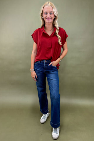 Woman wearing a red shirt, blue jeans, and white sneakers against a plain background