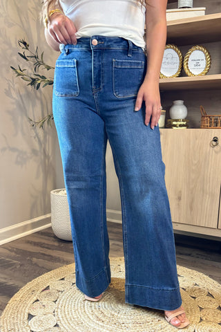 Close-up of a woman wearing high-waisted dark blue wide-leg jeans with large front patch pockets. She pairs them with a white ribbed tank top and nude open-toe block heels, standing on a woven circular rug with part of a potted plant and wooden shelving in the background.