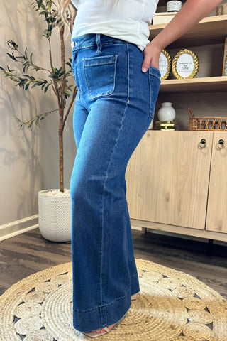 Close-up of a woman wearing high-waisted dark blue wide-leg jeans with large front patch pockets. She pairs them with a white ribbed tank top and nude open-toe block heels, standing on a woven circular rug with part of a potted plant and wooden shelving in the background.