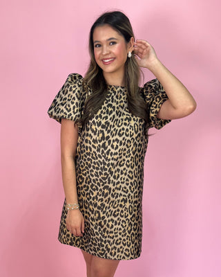 faux Leopard print mini dress with short flutter sleeves, a round neckline, and a relaxed shift silhouette, styled with gold strappy heels against a pink background.