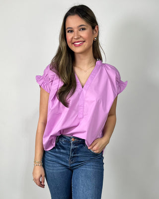 Woman wearing a bright lilac V-neck blouse with ruffle sleeves, paired with medium-wash cropped jeans and nude block-heel sandals, posing against a light gray backdrop.