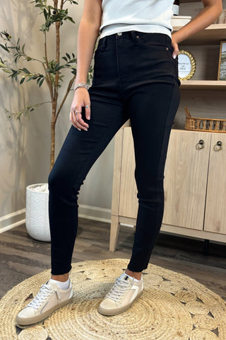 Close-up of a woman wearing high-waisted black skinny jeans paired with a white sleeveless knit top. She stands on a woven circular rug, wearing white sneakers, with part of a potted plant and wooden shelving visible in the background.