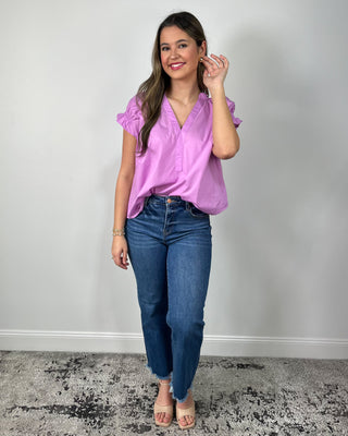Woman wearing a bright lilac V-neck blouse with ruffle sleeves, paired with medium-wash cropped jeans and nude block-heel sandals, posing against a light gray backdrop.