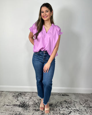 Woman wearing a bright lilac V-neck blouse with ruffle sleeves, paired with medium-wash cropped jeans and nude block-heel sandals, posing against a light gray backdrop.