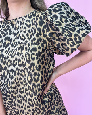 faux Leopard print mini dress with short flutter sleeves, a round neckline, and a relaxed shift silhouette, styled with gold strappy heels against a pink background.