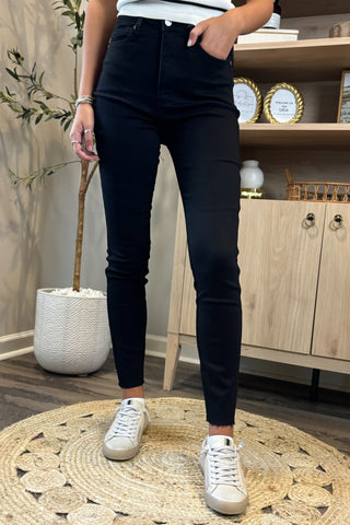 Close-up of a woman wearing high-waisted black skinny jeans paired with a white sleeveless knit top. She stands on a woven circular rug, wearing white sneakers, with part of a potted plant and wooden shelving visible in the background.