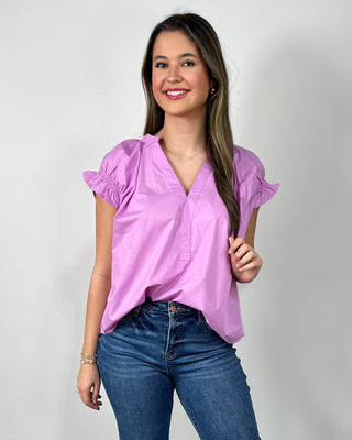 Woman wearing a bright lilac V-neck blouse with ruffle sleeves, paired with medium-wash cropped jeans and nude block-heel sandals, posing against a light gray backdrop.