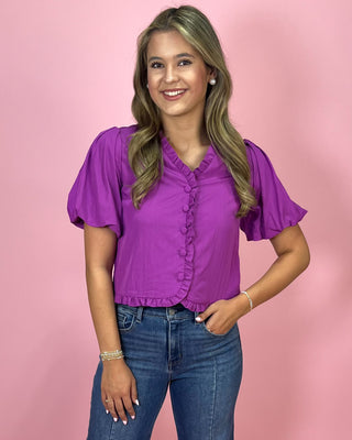Woman wearing a purple blouse with ruffled sleeves against a pink background