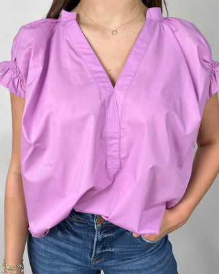 Woman wearing a bright lilac V-neck blouse with ruffle sleeves, paired with medium-wash cropped jeans and nude block-heel sandals, posing against a light gray backdrop.