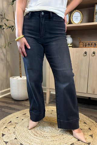 Close-up of a woman wearing high-waisted black wide-leg jeans paired with a white ribbed tank top. She stands on a woven circular rug, accessorized with nude pointed-toe heels, with part of a potted plant and wooden shelving in the background.
