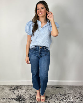 Woman wearing a light blue textured puff-sleeve blouse with a split V-neck, paired with medium-wash cropped jeans and nude block-heel sandals, posing against a light gray backdrop.
