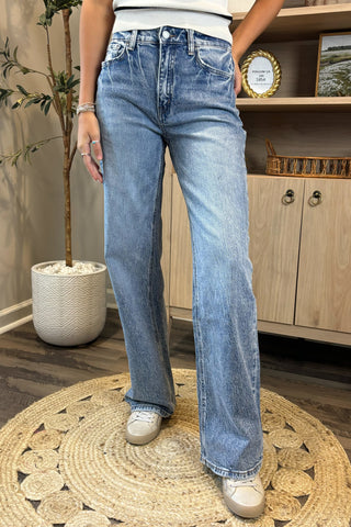 Close-up of a woman wearing high-waisted light blue wide-leg jeans with a slightly distressed and faded wash. She pairs them with a white sleeveless knit top and white sneakers, standing on a woven circular rug with a potted plant and wooden shelving in the background.