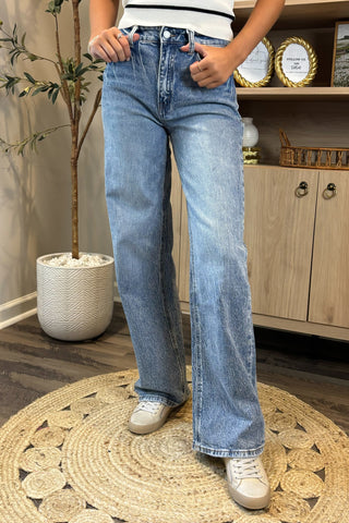 Close-up of a woman wearing high-waisted light blue wide-leg jeans with a slightly distressed and faded wash. She pairs them with a white sleeveless knit top and white sneakers, standing on a woven circular rug with a potted plant and wooden shelving in the background.