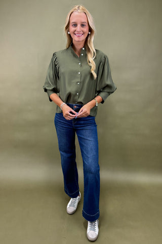 Woman wearing an olive green blouse and blue jeans against a beige background
