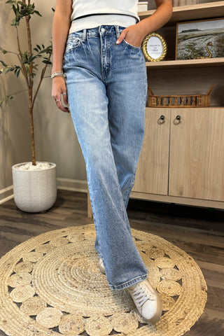 Close-up of a woman wearing high-waisted light blue wide-leg jeans with a slightly distressed and faded wash. She pairs them with a white sleeveless knit top and white sneakers, standing on a woven circular rug with a potted plant and wooden shelving in the background.