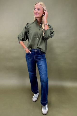Woman wearing a green shirt, blue jeans, and white sneakers against a plain background