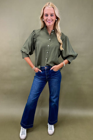 Woman wearing an olive green blouse and blue jeans against a beige background