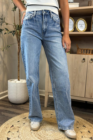 Close-up of a woman wearing high-waisted light blue wide-leg jeans with a slightly distressed and faded wash. She pairs them with a white sleeveless knit top and white sneakers, standing on a woven circular rug with a potted plant and wooden shelving in the background.