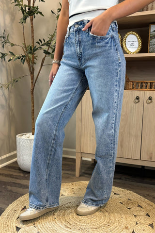 Close-up of a woman wearing high-waisted light blue wide-leg jeans with a slightly distressed and faded wash. She pairs them with a white sleeveless knit top and white sneakers, standing on a woven circular rug with a potted plant and wooden shelving in the background.