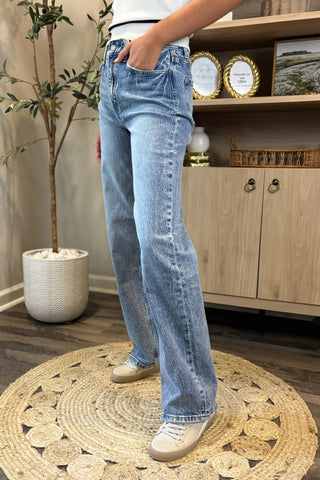 Close-up of a woman wearing high-waisted light blue wide-leg jeans with a slightly distressed and faded wash. She pairs them with a white sleeveless knit top and white sneakers, standing on a woven circular rug with a potted plant and wooden shelving in the background.