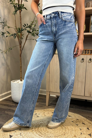 Close-up of a woman wearing high-waisted light blue wide-leg jeans with a slightly distressed and faded wash. She pairs them with a white sleeveless knit top and white sneakers, standing on a woven circular rug with a potted plant and wooden shelving in the background.