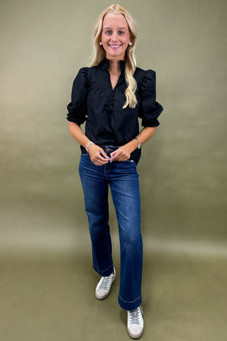 Woman wearing a black blouse, blue jeans, and white sneakers against a beige background