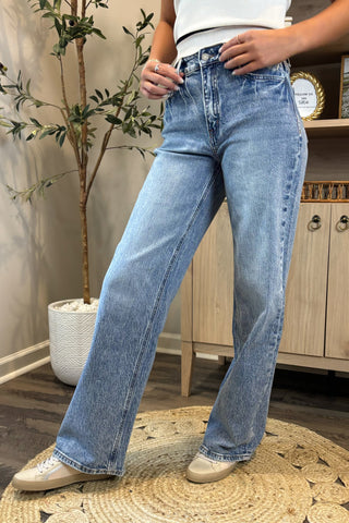 Close-up of a woman wearing high-waisted light blue wide-leg jeans with a slightly distressed and faded wash. She pairs them with a white sleeveless knit top and white sneakers, standing on a woven circular rug with a potted plant and wooden shelving in the background.