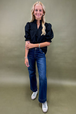 Woman wearing a black blouse, blue jeans, and white sneakers against a beige background