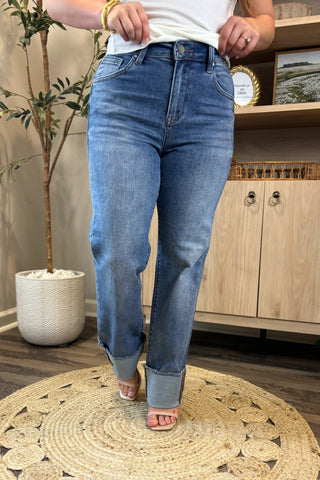 Close-up of a woman wearing high-waisted medium-wash jeans with wide, folded cuffs and raw hems. She pairs them with a white ribbed tank top and nude block-heel sandals, standing on a woven circular rug with a potted plant and wooden shelving in the background.