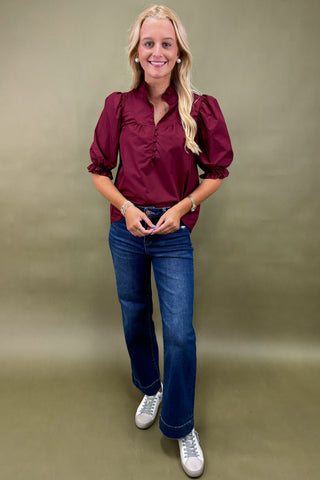 Woman wearing a burgundy blouse and blue jeans against a plain background