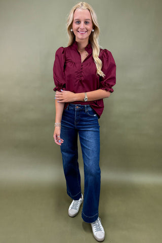 Woman wearing a burgundy blouse, blue jeans, and white sneakers against a green background