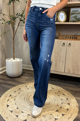Close-up of a woman wearing high-waisted dark blue jeans with light distressing near the pockets and thigh. She pairs them with a white sleeveless knit top and white sneakers, standing on a woven circular rug with a potted plant and wooden shelving in the background.