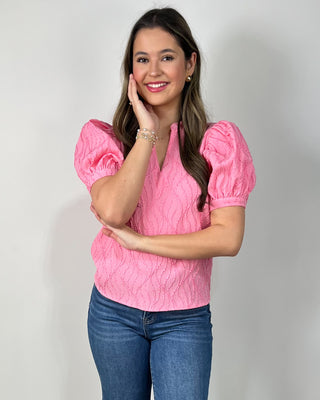 Woman wearing a textured hot pink puff-sleeve blouse with a V-neck, paired with medium-wash cropped jeans and nude block-heel sandals, posing against a light gray backdrop.