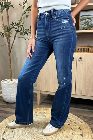 Close-up of a woman wearing high-waisted dark blue jeans with light distressing near the pockets and thigh. She pairs them with a white sleeveless knit top and white sneakers, standing on a woven circular rug with a potted plant and wooden shelving in the background.