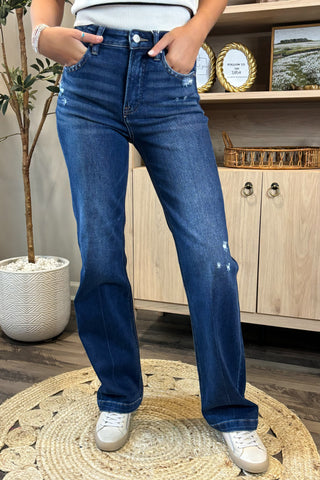 Close-up of a woman wearing high-waisted dark blue jeans with light distressing near the pockets and thigh. She pairs them with a white sleeveless knit top and white sneakers, standing on a woven circular rug with a potted plant and wooden shelving in the background.