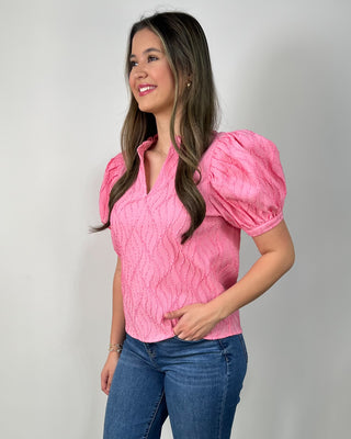 Woman wearing a textured hot pink puff-sleeve blouse with a V-neck, paired with medium-wash cropped jeans and nude block-heel sandals, posing against a light gray backdrop.