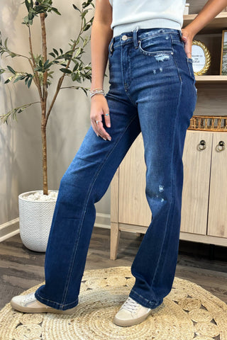 Close-up of a woman wearing high-waisted dark blue jeans with light distressing near the pockets and thigh. She pairs them with a white sleeveless knit top and white sneakers, standing on a woven circular rug with a potted plant and wooden shelving in the background.