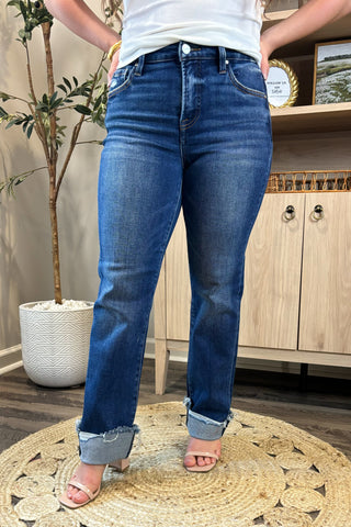 Close-up of a woman wearing high-waisted dark blue straight-leg jeans with distressed, rolled cuffs. She pairs them with a white ribbed tank top and nude block-heel sandals, standing on a woven circular rug with a potted plant and wooden shelving in the background.