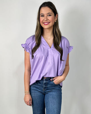 Woman wearing a lavender ruffle-sleeve blouse with a V-neckline, styled with medium-wash cropped jeans and nude block-heel sandals, posing against a light gray backdrop.