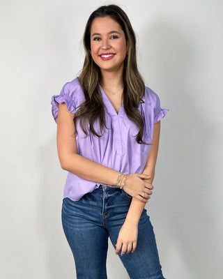 Woman wearing a lavender ruffle-sleeve blouse with a V-neckline, styled with medium-wash cropped jeans and nude block-heel sandals, posing against a light gray backdrop.