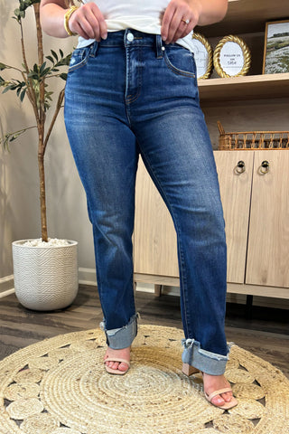 Close-up of a woman wearing high-waisted dark blue straight-leg jeans with distressed, rolled cuffs. She pairs them with a white ribbed tank top and nude block-heel sandals, standing on a woven circular rug with a potted plant and wooden shelving in the background.