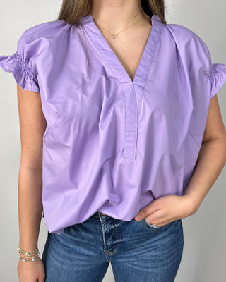 Woman wearing a lavender ruffle-sleeve blouse with a V-neckline, styled with medium-wash cropped jeans and nude block-heel sandals, posing against a light gray backdrop.