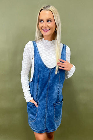 Woman wearing a blue denim dress over a white turtleneck against a green background