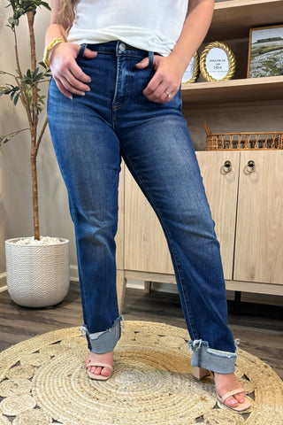 Close-up of a woman wearing high-waisted dark blue straight-leg jeans with distressed, rolled cuffs. She pairs them with a white ribbed tank top and nude block-heel sandals, standing on a woven circular rug with a potted plant and wooden shelving in the background.