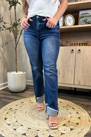 Close-up of a woman wearing high-waisted dark blue straight-leg jeans with distressed, rolled cuffs. She pairs them with a white ribbed tank top and nude block-heel sandals, standing on a woven circular rug with a potted plant and wooden shelving in the background.
