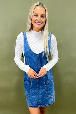 Woman wearing a denim dress over a white long-sleeve shirt against a green background