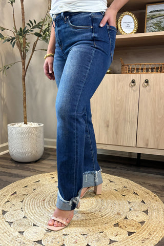Close-up of a woman wearing high-waisted dark blue straight-leg jeans with distressed, rolled cuffs. She pairs them with a white ribbed tank top and nude block-heel sandals, standing on a woven circular rug with a potted plant and wooden shelving in the background.
